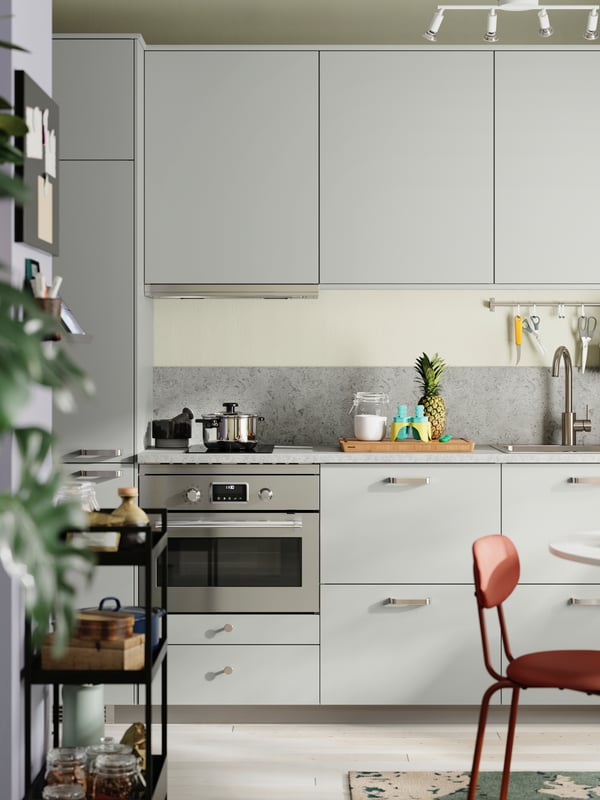 A kitchen with light grey HAVSTORP fronts has an oven, an induction hob with a pot on it, and a KUNGFORS rail with hooks.