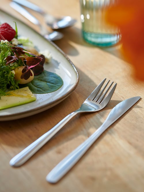 A single GLADELIG plate with a fresh salad, FÖRNUFT cutlery on the side, all placed on a light wooden surface.