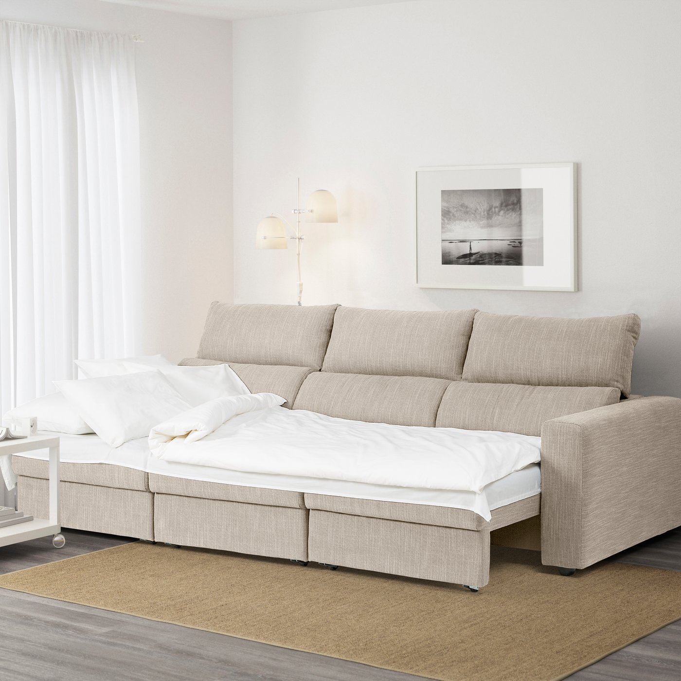 All'interno di un soggiorno chiaro con pareti e tende bianche, è presente un divano letto ESKILSTUNA a 3 posti in tessuto beige aperto in versione letto matrimoniale.