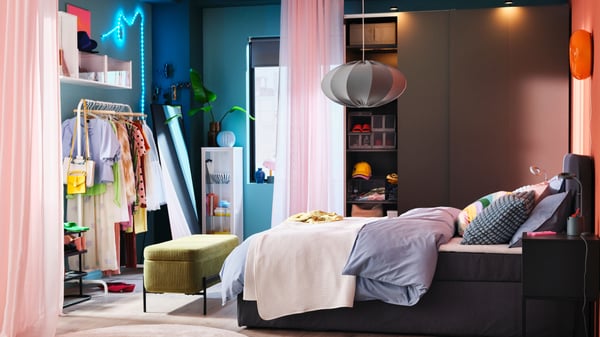 A colourful bedroom with a BLÅFJÄLLET ottoman divan bed in front of a dark grey PAX/HASVIK wardrobe and a pink curtain.