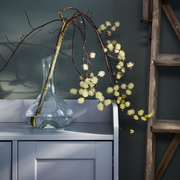 A lone, blooming, sculptural branch in a STORSINT clear glass carafe, in turn is standing on top of a grey HAUGA sideboard.