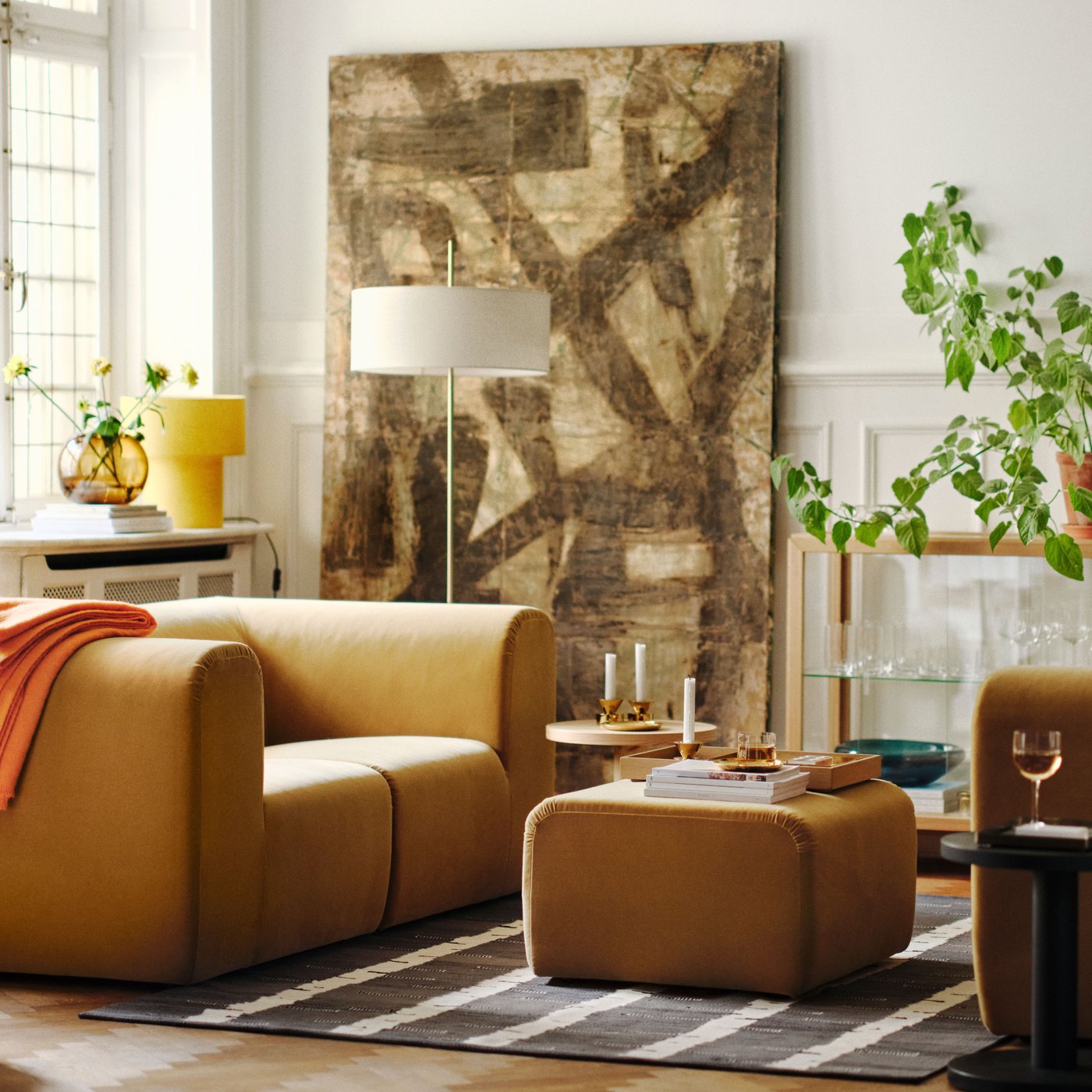 Es ist ein helles Wohnzimmer mit zwei beigefarbenen STOCKHOLM 2025 2er-Sofas zu sehen, die sich gegenüberstehen. Dazwischen steht ein Hocker auf einem Teppich.