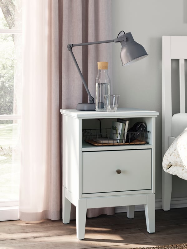 A dark desk lamp standing on a white IDANÄS bedside table next to a white IDANÄS double bed in a dimly sunlit, light bedroom.