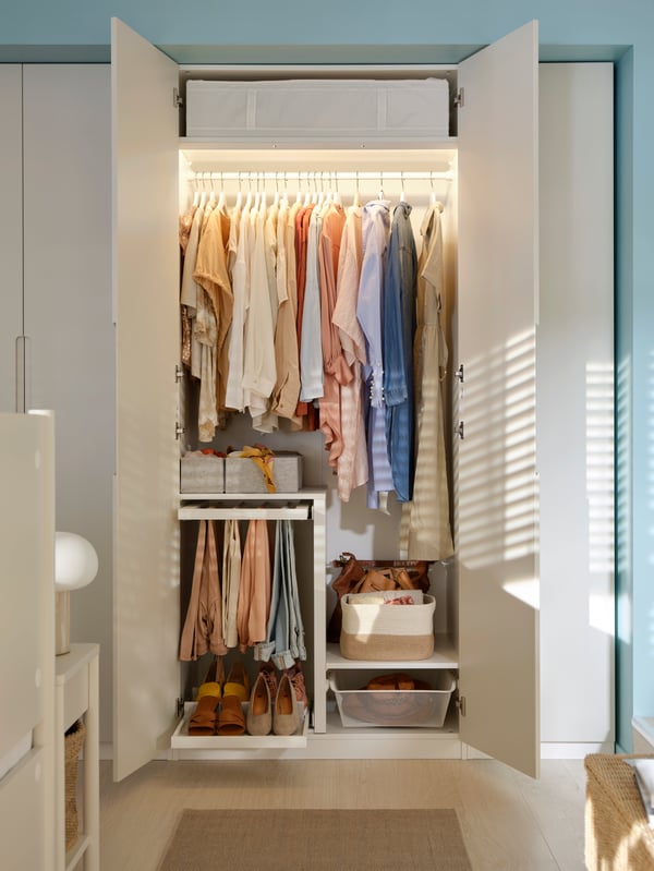A white PAX wardrobe with off-white TONSTAD doors and wardrobe lighting filled with neatly arranged clothes, shoes, and more.