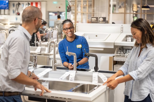 An IKEA co-worker in a blue striped shirt is smiling and talking to two people in-store and showing sinks and faucets.