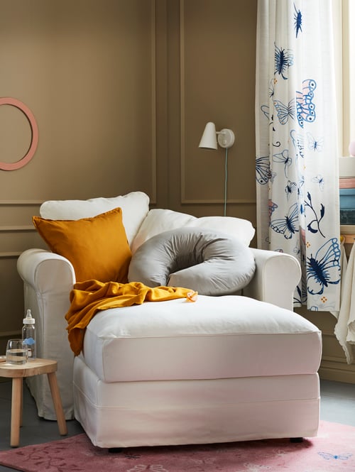 A chaise longue is in a corner and has a few cushions on it, including a LEN grey nursing pillow that is half-circle shaped.
