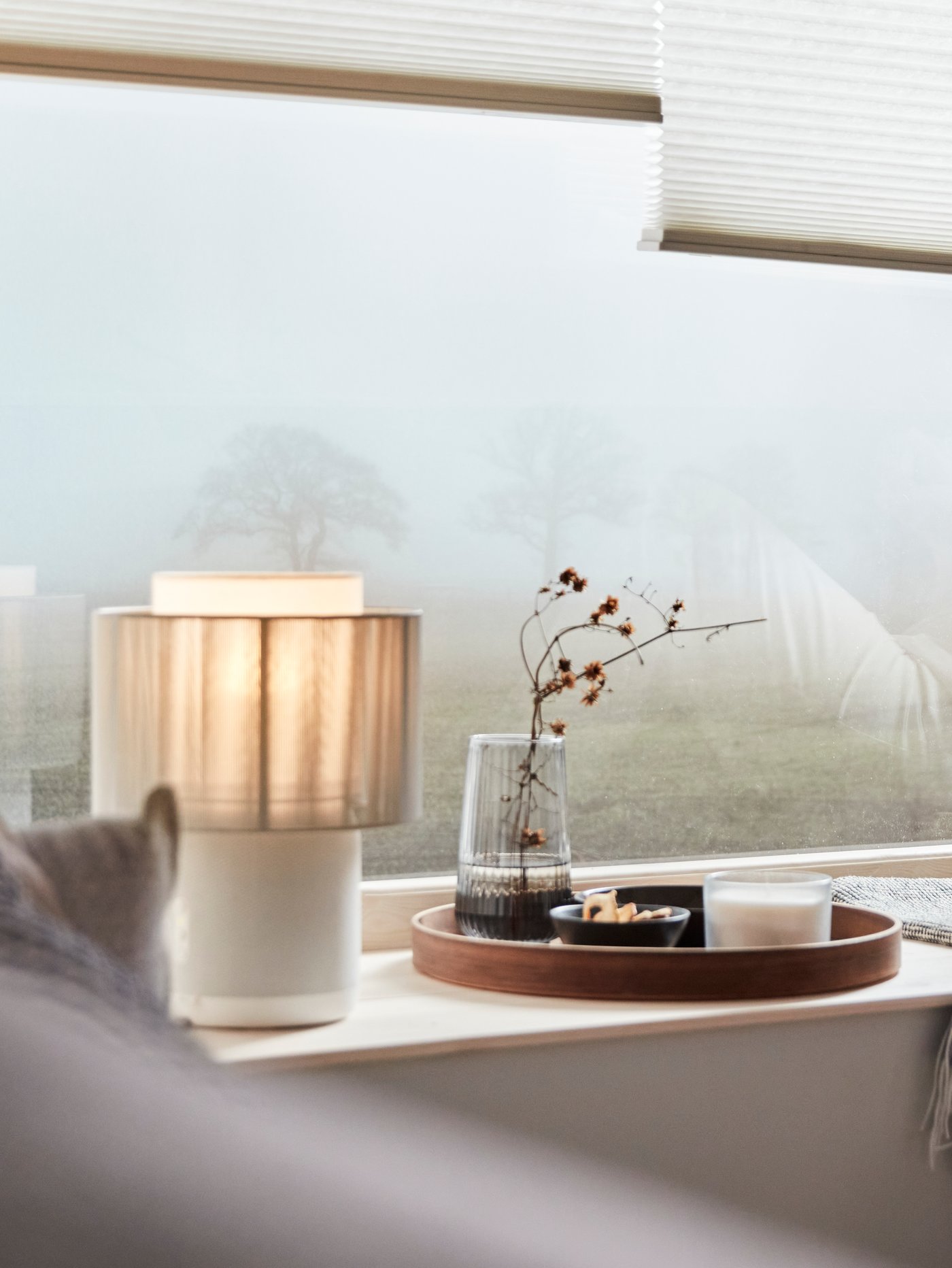 A SYMFONISK speaker lamp with textile shade on a window sill. Outside there is a tree, barely visible through the mist.