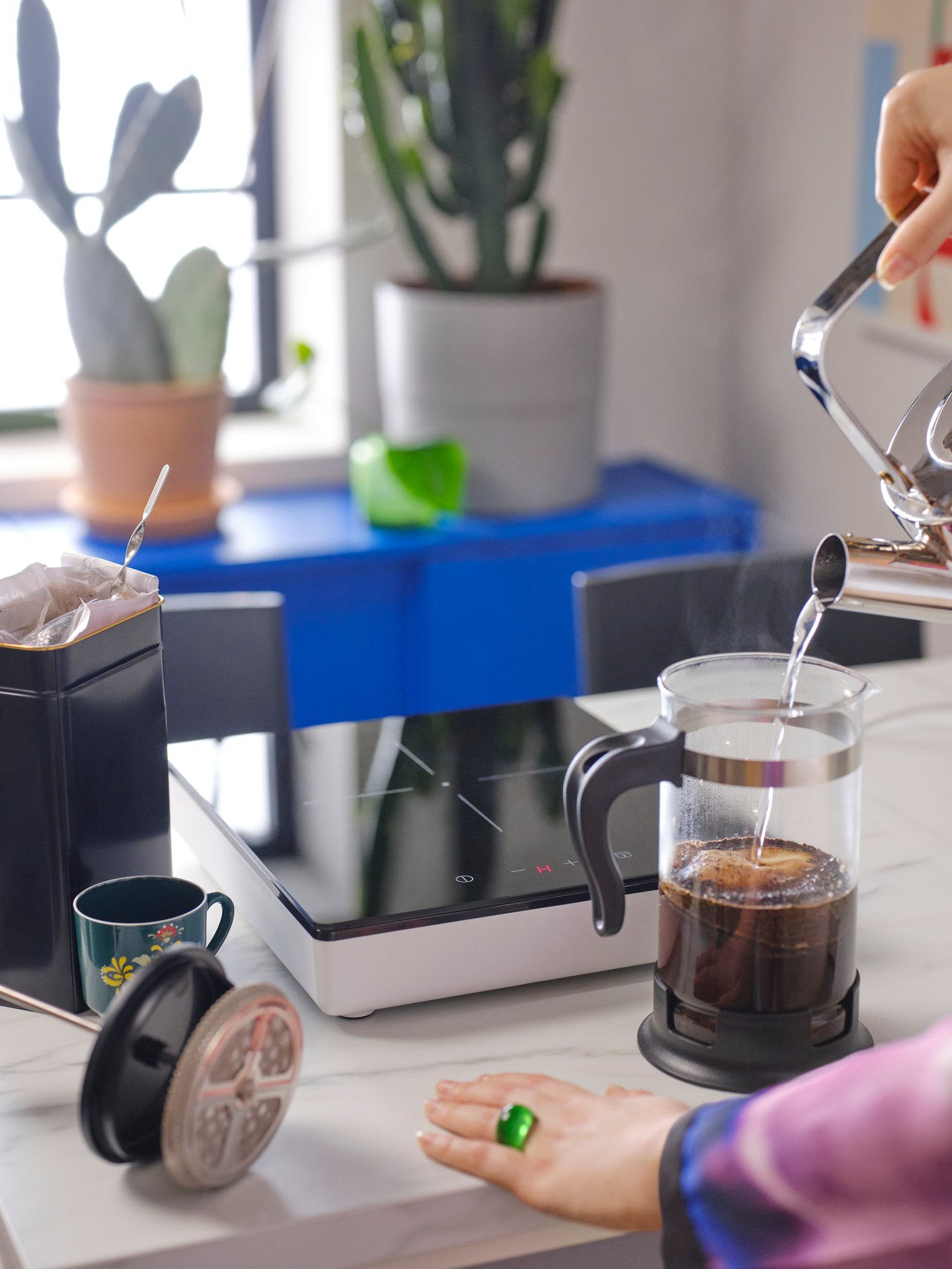 A hand by an UPPHETTA coffee/tea maker in glass and stainless steel and a TILLREDA portable induction hob 1 zone in white.