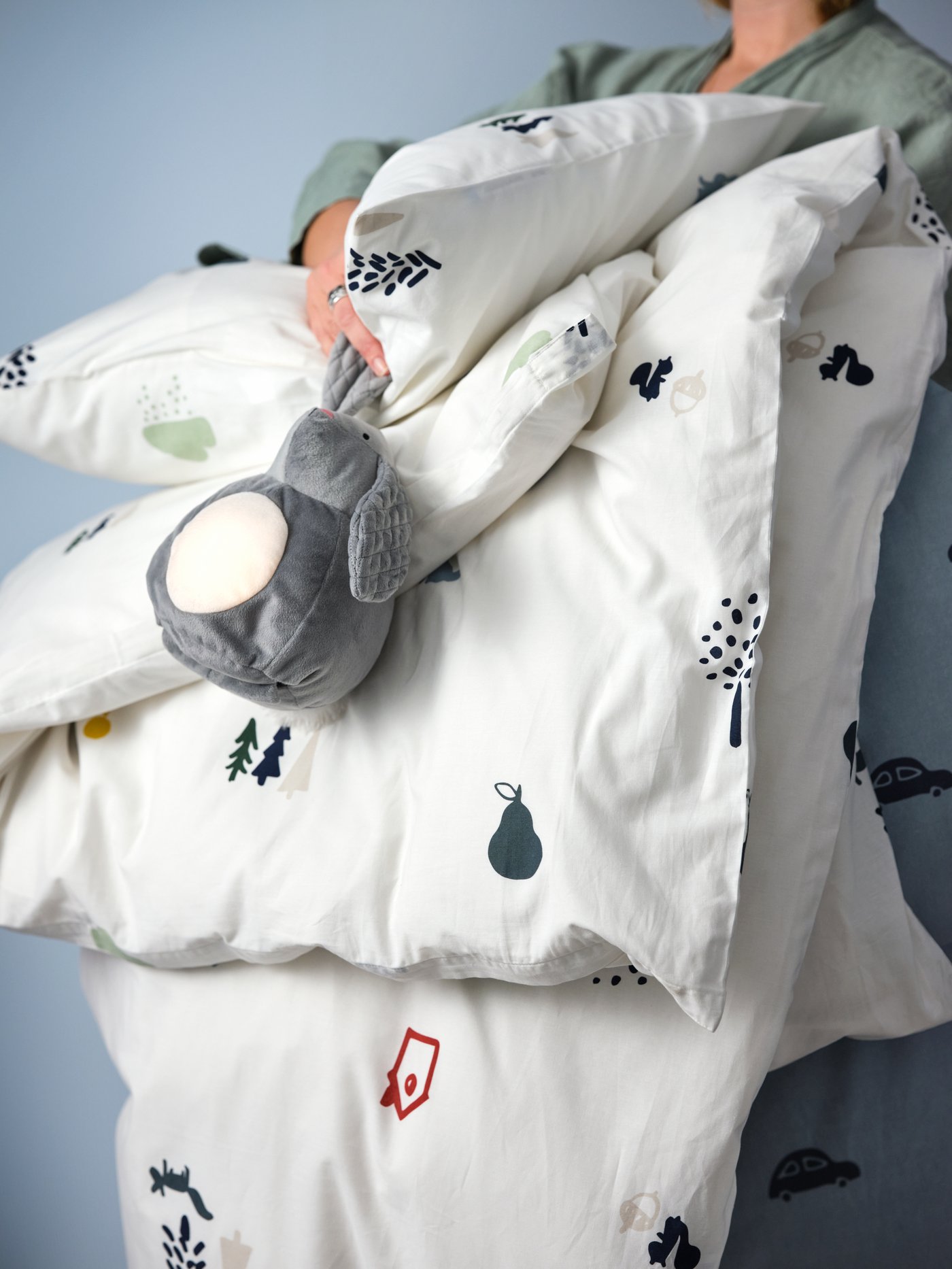 Osoba drží složenou bílou dětskou peřinu s barevnými potisky stromů, zvířat a ovoce. Na peřině je šedá plyšová hračka s velkýma ušima.