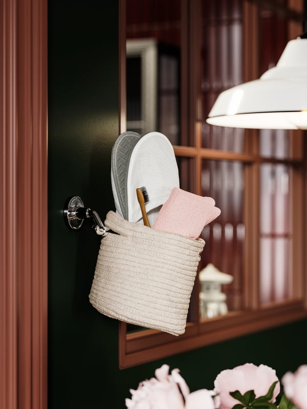 A TJABBIG basket hanging on a wall hook, holding white slippers and a pink towel in a bathroom with dark green walls.