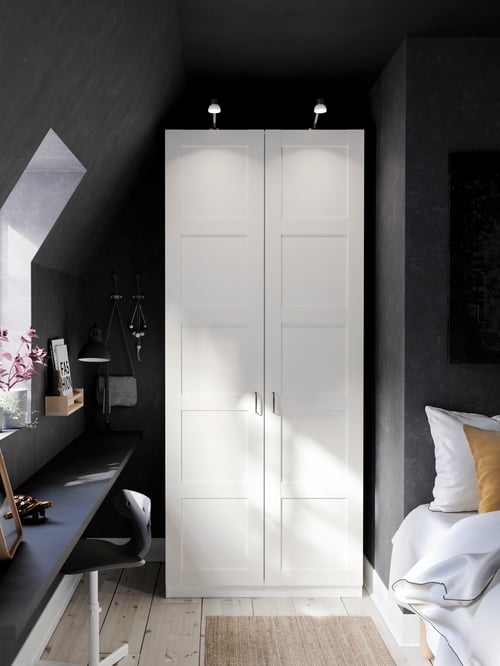 A grey room with a sloped ceiling has a closed PAX wardrobe with Bergsbo white hinged doors in a nook by a desk and a bed.