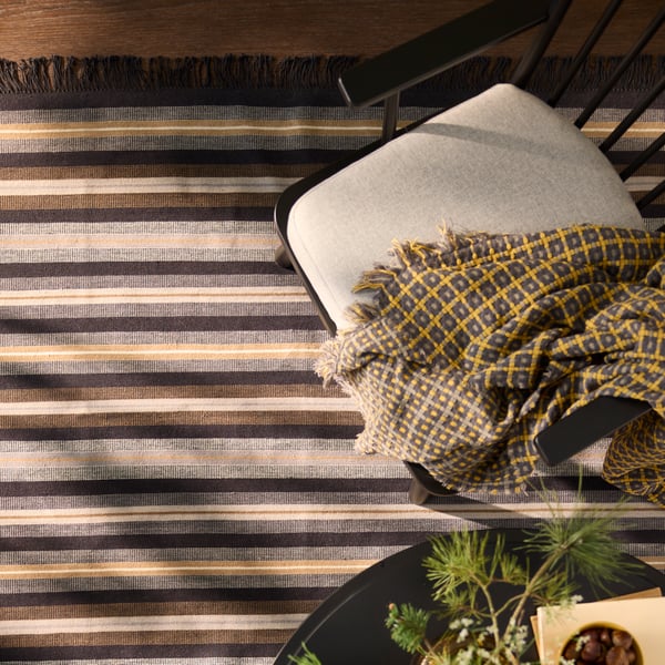 A flatwoven black/brown and grey RÅVAROR rug is elegantly placed on a wooden floor beside a chair and patterned throw.
