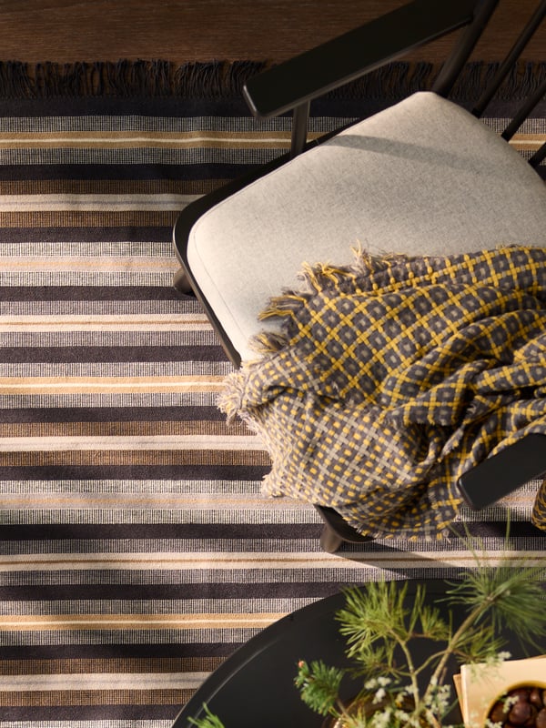 A flatwoven black/brown and grey RÅVAROR rug is elegantly placed on a wooden floor beside a chair and patterned throw.