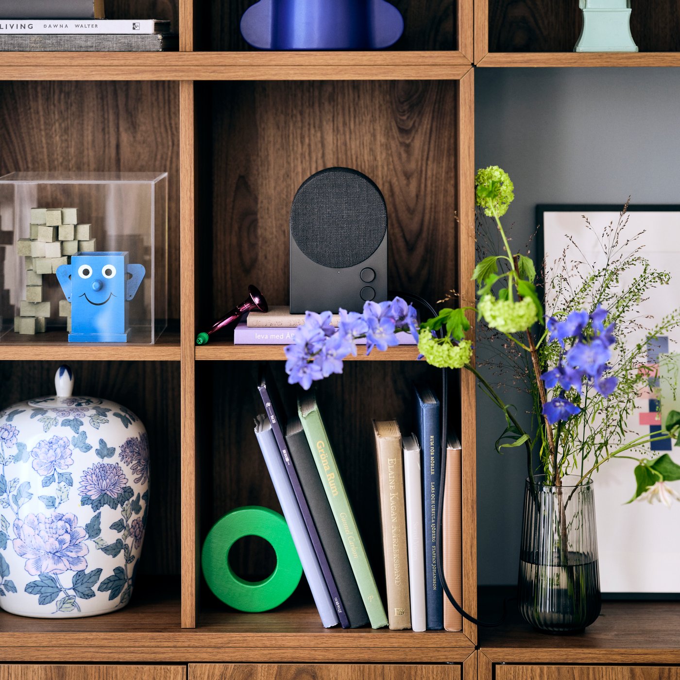 A storage unit with shelves holding a NATTBAD Bluetooth speaker, a number of books, a ceramic pot and a painting in a frame.