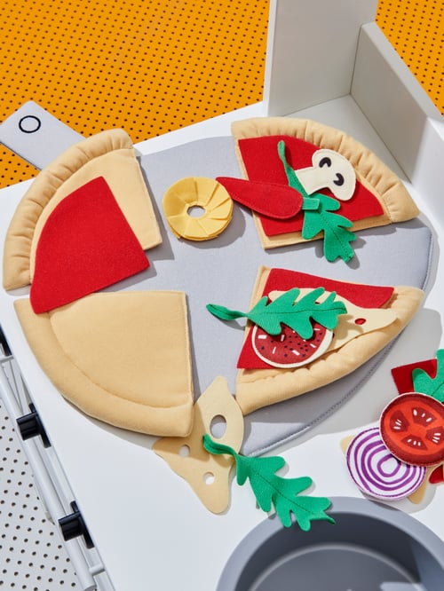 Prin-plan cu pizza de jucărie DUKTIG din țesătură moale, cu topping-uri divers colorate, pe un cuptor de jucărie alb pentru copii.