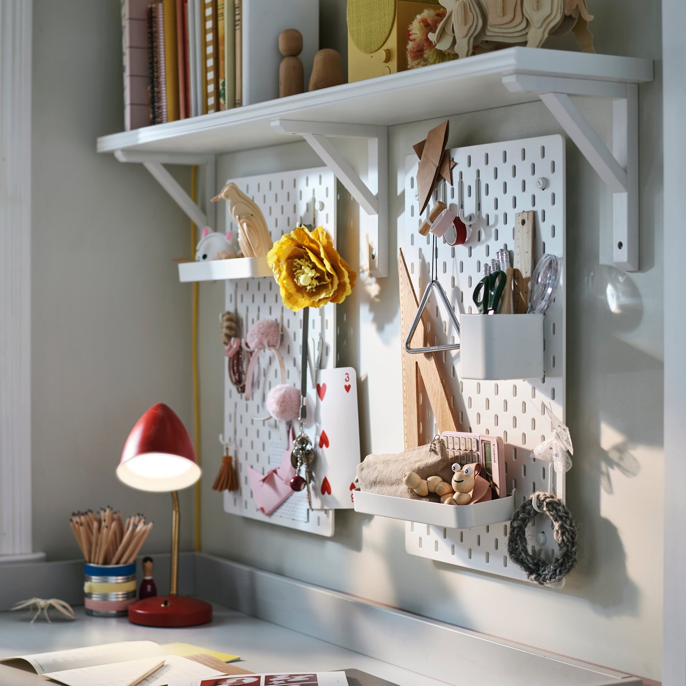 Deux combinaisons de panneaux perforés SKÅDIS blancs fixés au mur accueillent les fournitures et accessoires, et une lampe de bureau à LED ISNÅLEN rouge est posée à proximité.