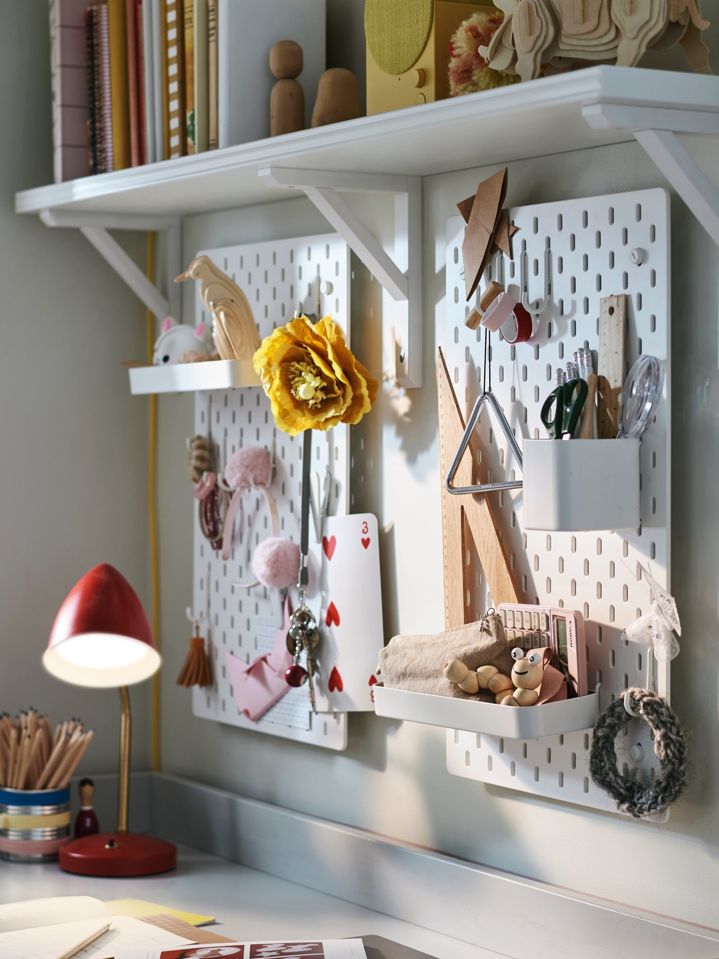Two white SKÅDIS pegboard combinations on a wall hold stationery and accessories with a red ISNÅLEN LED work lamp nearby.