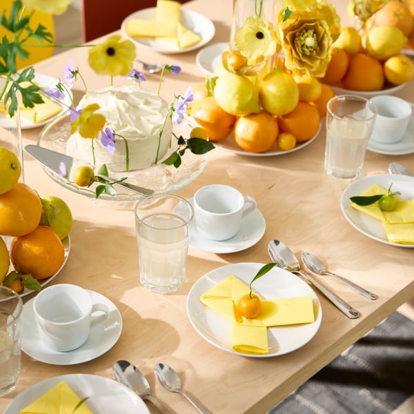 A birthday table setting has white plates with yellow paper napkins, POKAL glasses and plates with oranges and lemons.