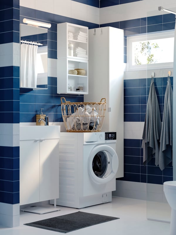 Un bagno con mobili bianchi e una lavatrice. Le pareti hanno piastrelle bianche e blu. A lato, a dei ganci sono appesi con degli asciugamani.