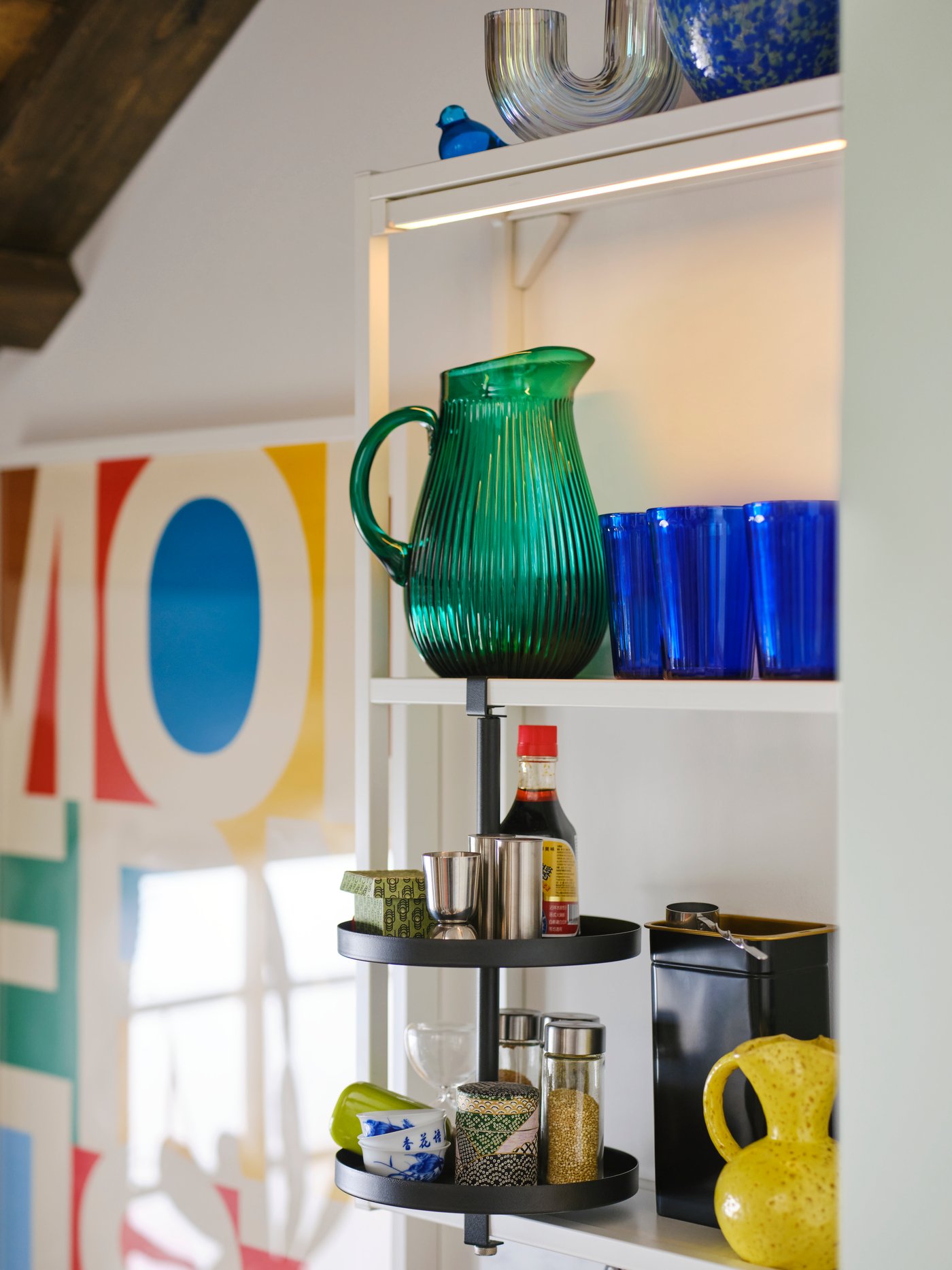 An ENHET wall frame with shelves in white holding a SÄLLSKAPLIG jug 2L patterned green and VARDAGEN glasses in blue.