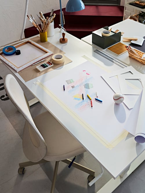 Workspace with a beige office chair, two white desks, pieces of paper, drawing utensils, paint brushes, a blue desk lamp.
