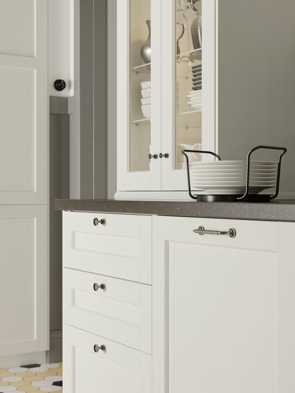 A cabinet with white ENKÖPING glass doors on a countertop, holding bowls, plates and jugs, with a plate holder on the side.