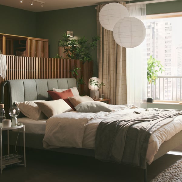 Ein gepolstertes TÄLLÅSEN Bettgestell in Graugrün mit einem Sichtschutz hinter dem Bettgestell steht in einem ruhigen grünen Schlafzimmer mit grossen Fenstern.
