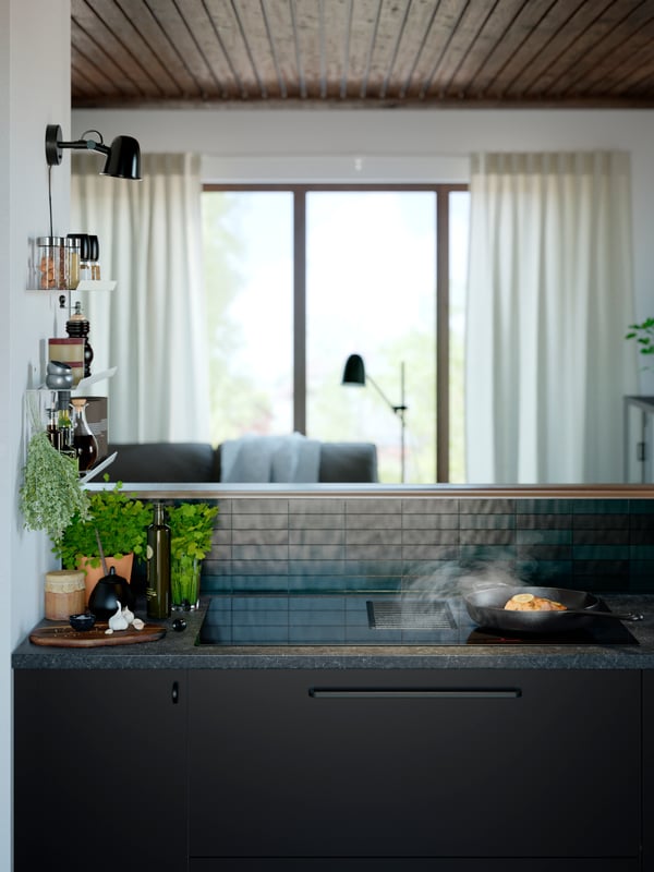 Food is being prepared in a cast iron frying pan on a FÖRNEBO induction cooktop next to a pot of herbs and bottle of oil.