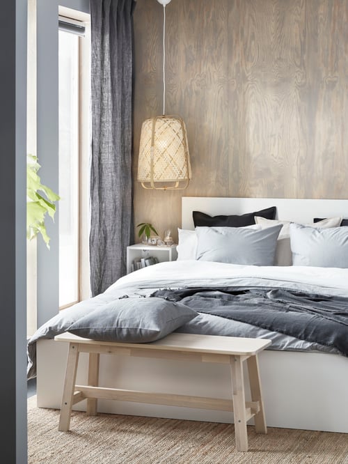 A light-filled bedroom with a wood wall, windows and a MALM high white bed frame with white and grey bedding on a jute rug.
