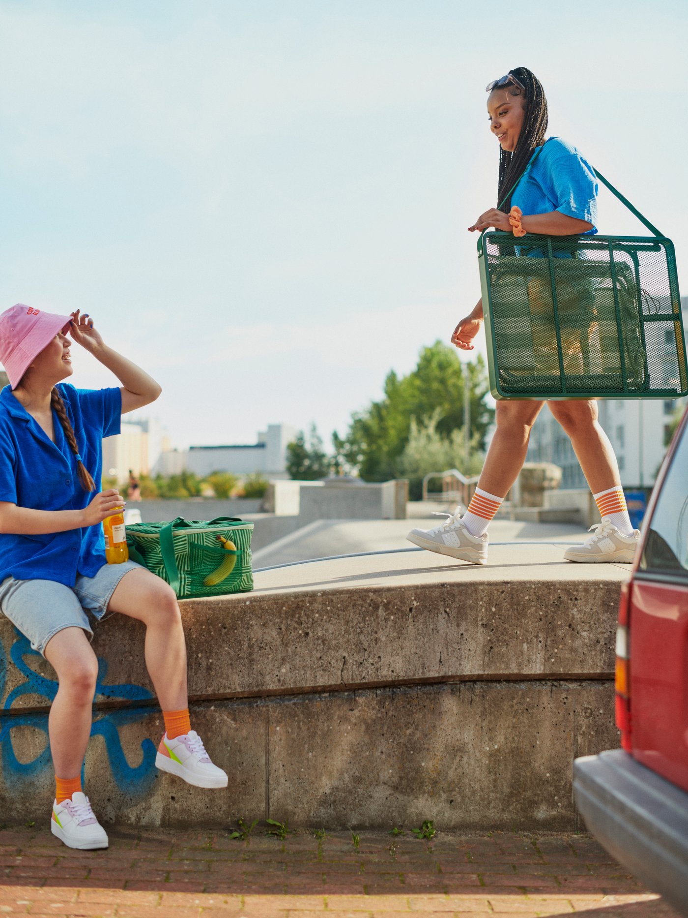 Jeunes personnes dans un paysage urbain ensoleillé. L’une d’entre elles porte un ensemble table et tabouret pliants STRANDÖN, l’autre est assise à côté d’un sac isotherme NÄBBFISK.