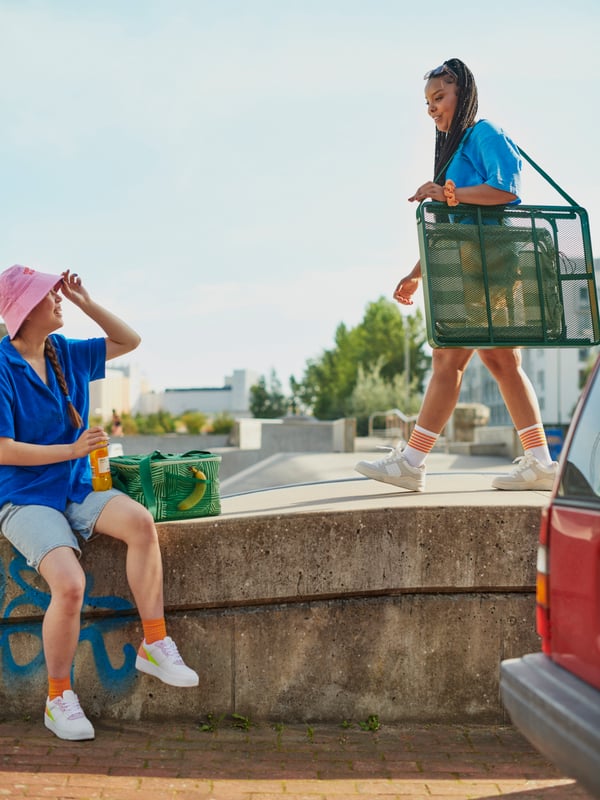 Young people in a sunny city landscape. One carries a STRANDÖN folding table set, one sits next to a NÄBBFISK cooling bag.