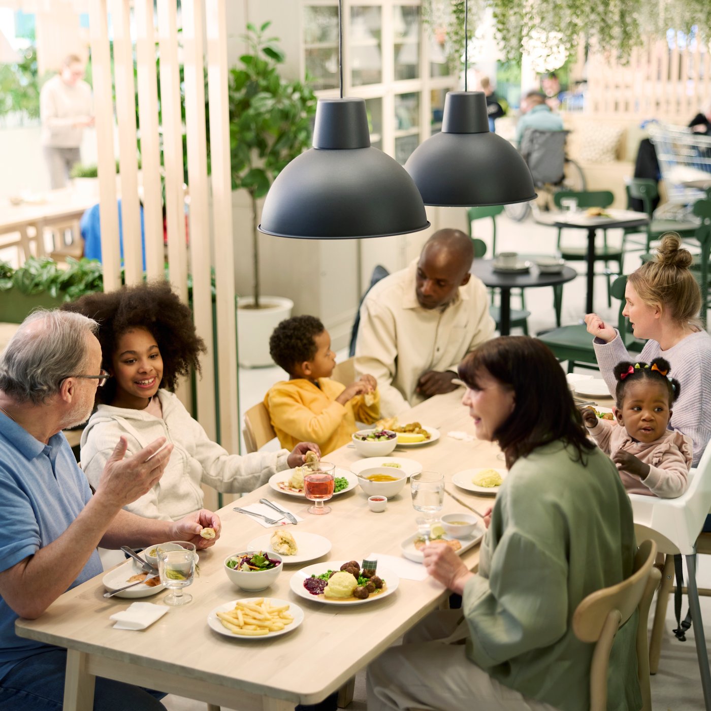 Eine Gruppe von Menschen unterschiedlichen Alters sitzt gemeinsam an einem Esstisch im IKEA Restaurant und genießt eine Mahlzeit.