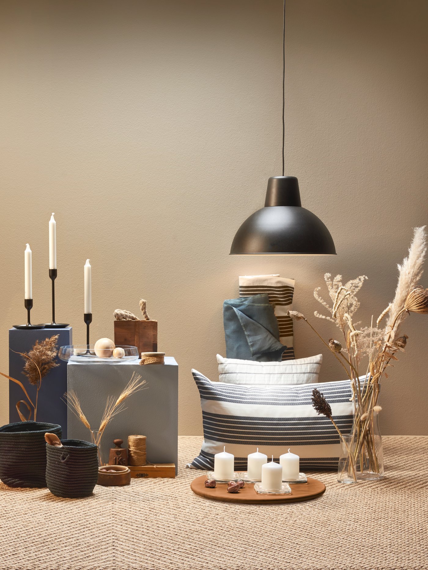 Zona del salón con una lámpara colgante negra, velas en bloque sin aroma, candelabros, cojines, un jarrón y cestas.
