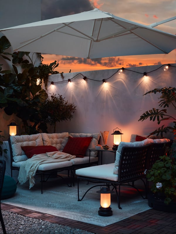 HÖGÖN parasol danner ramme omkring et loungeområde med JUTHOLMEN loungemøbler i en aftenstund, hvor SVARTRÅ lyskæde og STORHAGA lampe lyser op.