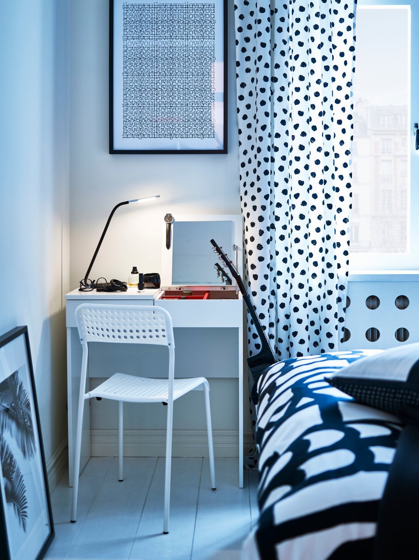 Zona de dormitorio con un tocador blanco con lámpara de trabajo y silla, con una cama a un lado y un marco en la pared de arriba.