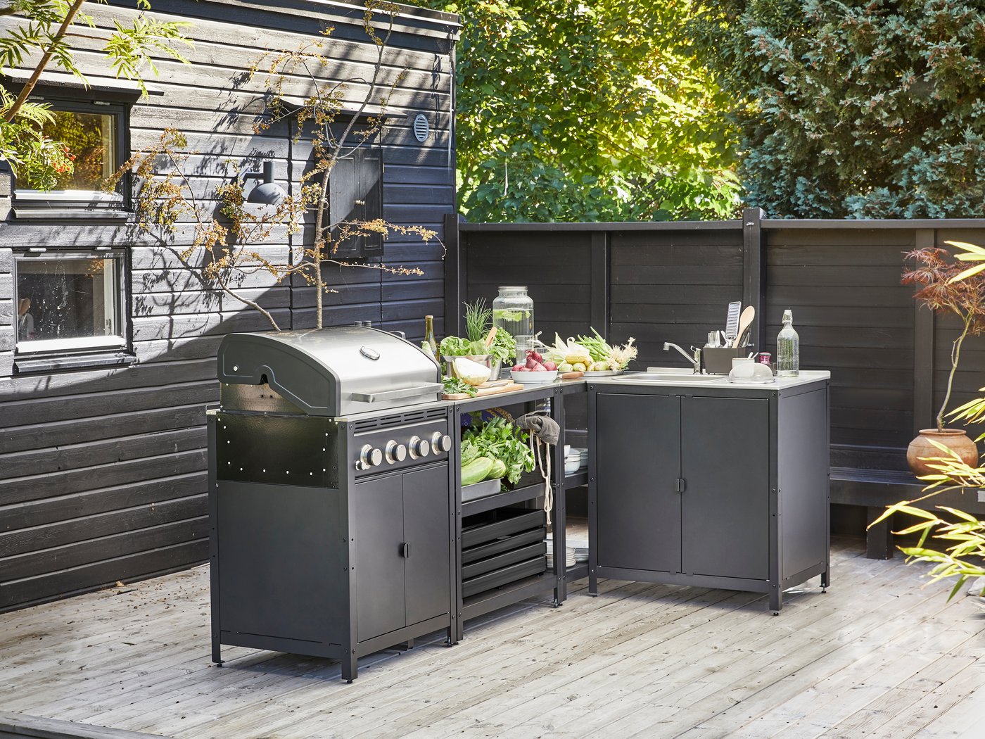 Een stijlvolle IKEA GRILLSKÄR buitenkeukenopstelling, bestaande uit een gasbarbecue, een voorbereidingseenheid gevuld met verse groenten en een spoelbakmodule, staat op een houten terras naast een donkerhouten huis, klaar voor een buitenmaaltijd.