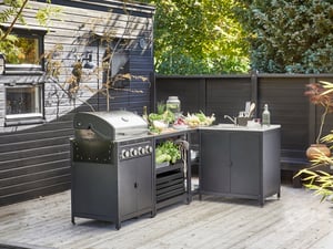 An undefinedA stylish IKEA GRILLSKÄR outdoor kitchen setup, featuring a gas barbecue, a preparation unit filled with fresh vegetables, and a sink module, sits on a wooden deck next to a dark-paneled house, ready for an outdoor me