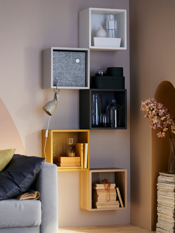 Un pezzo di divano grigio accanto a una parete con appeso mobili EKET gialli, grigi e una nera.  All'interno dei mobili da parete ci sono libri, una cassa wi-fi e contenitori on vetro. 