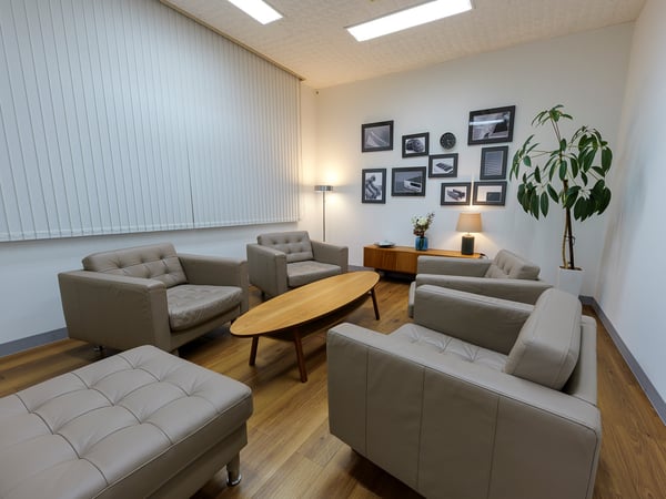 ベージュのソファと木製ローテーブルを置いた応接スペース。壁に多数のフレームと観葉植物があり、間接照明が柔らかく照らしている。