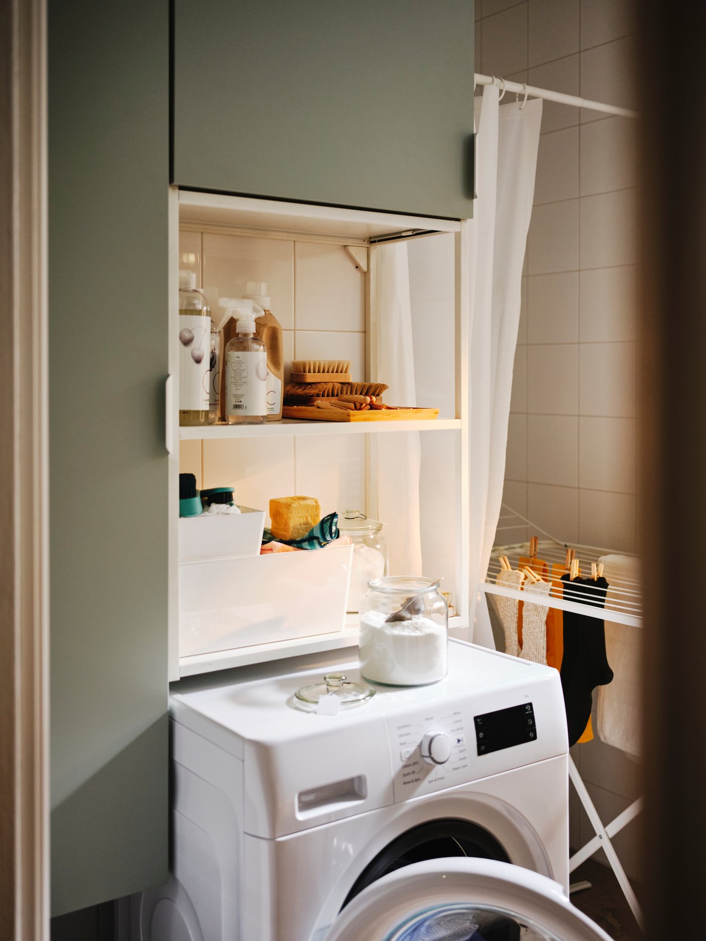 Pračka IKEA 300 UDDARP pod bílo-světle šedozelenou úložnou sestavou ENHET uzpůsobenou na praní prádla.