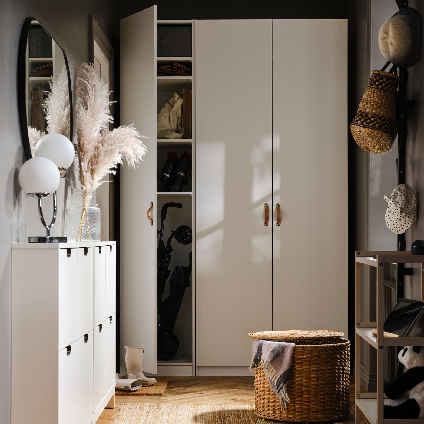 En hall med vita förvaringsskåp, spegel och bordslampa, med korgar, hattar och en stor garderob längst in samt en rund korg på golvet.