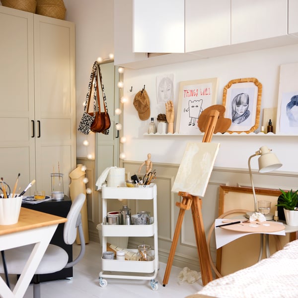 A creative crafting area featuring a white RÅSKOG trolley, holding art supplies next to a desk and an easel in a bright room.