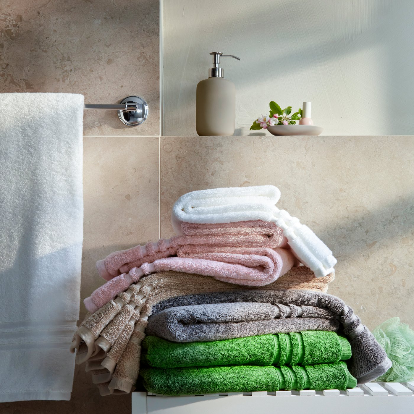 A stack of different coloured, 100% cotton towels, including BBROKGLIM white bath towel, are on a white bench by a tiled wall.