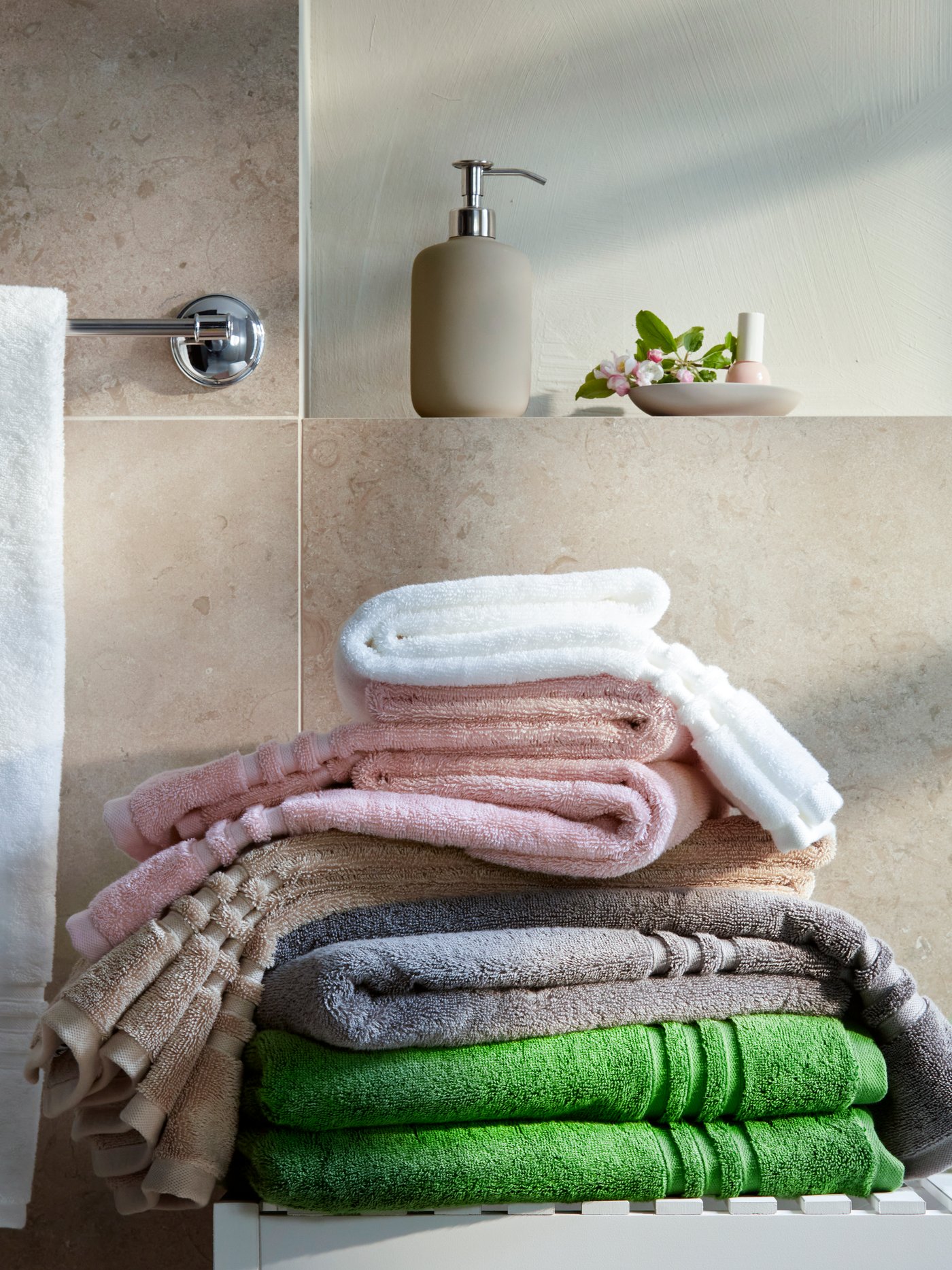 Auf einer weißen Bank an einer gefliesten Wand liegt ein Stapel verschiedenfarbiger Handtücher aus 100% Baumwolle, darunter auch ein weißes BBROKGLIM Badetuch.
