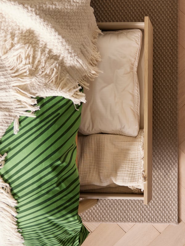 A LAPPTÅG throw lies on a TONSTAD bed with storage. In one of the bed’s drawers, there is a cushion and a VALLKRASSING throw.