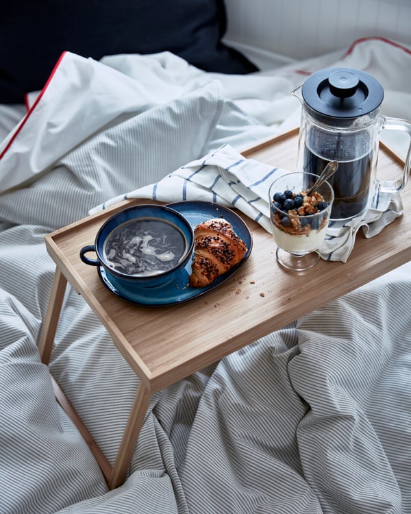 Sängyssä RESGODS-vuodetarjotin, jolle katettu kahvia, croissant ja jogurttia sekä marjoja jalkalasiin.