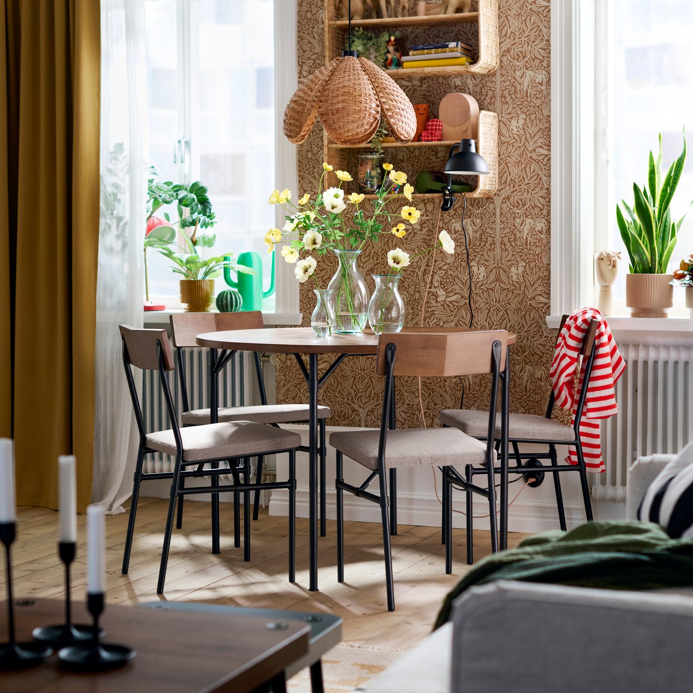 A dining space with four GRÖTÅN chairs in Tibbleby dark beige, placed around a wooden table with flowers and shelves above.