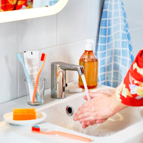 Person washing hands with a BROGRUND wash-basin mixer tap with sensor, surrounded by colourful toothbrushes and soap.