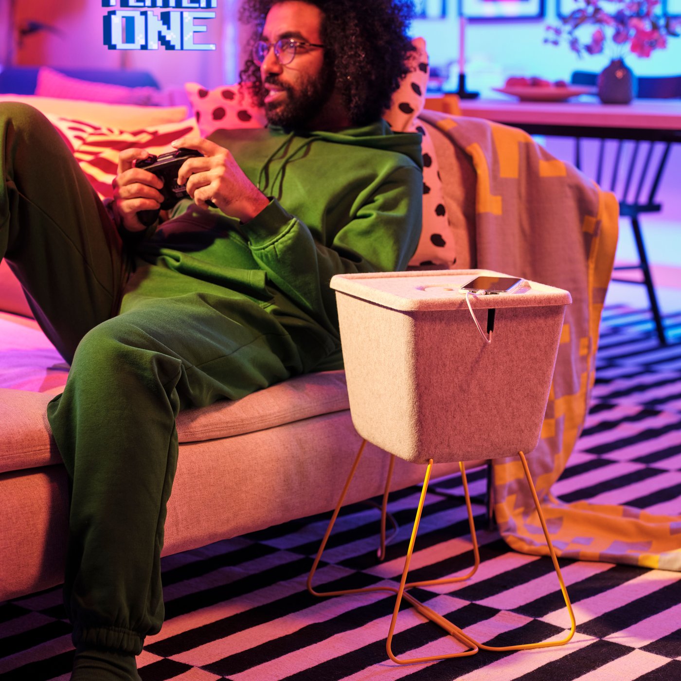 Mężczyzna grający na konsoli na sofie. Obok niego stoi kosz BRÄNNBOLL.