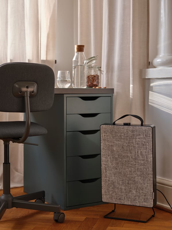 A black FÖRNUFTIG air purifier standing on the floor beneath a study space with a dark grey BLECKBERGET swivel chair.